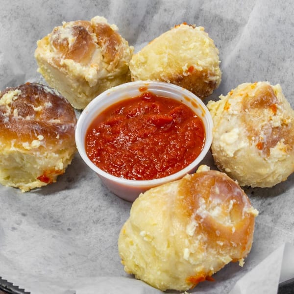 Five golden-brown garlic knots with cheese and a side of red marinara sauce in a white cup.